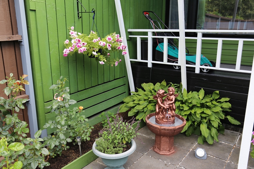 Portin vieressä kasvaa alppiruusupensaan lisäksi vorenkilpiä sekä ryhmässä punakärsämöitä ja yksivuotisia kesäsäsdekukkia. Valkoinen koristekate tuo valoa nurkkaan.