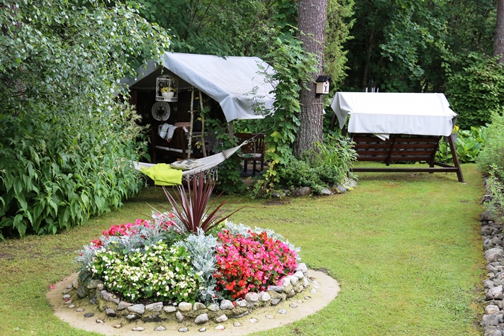 Pihalla on useita kukka-asetelmia, tässä kukkivat kesäbegoniat keskellään punalehtinen verililjapuu (Cordyline) ja väleissään hopeavillakkoa. Taustalla on Hönölä. 